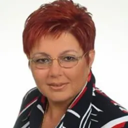 Portret kobiety w okularach, z krótkimi rudymi włosami, ubranej w marynarkę w kolorach granatu, bieli i czerwieni, na jasnym tle.