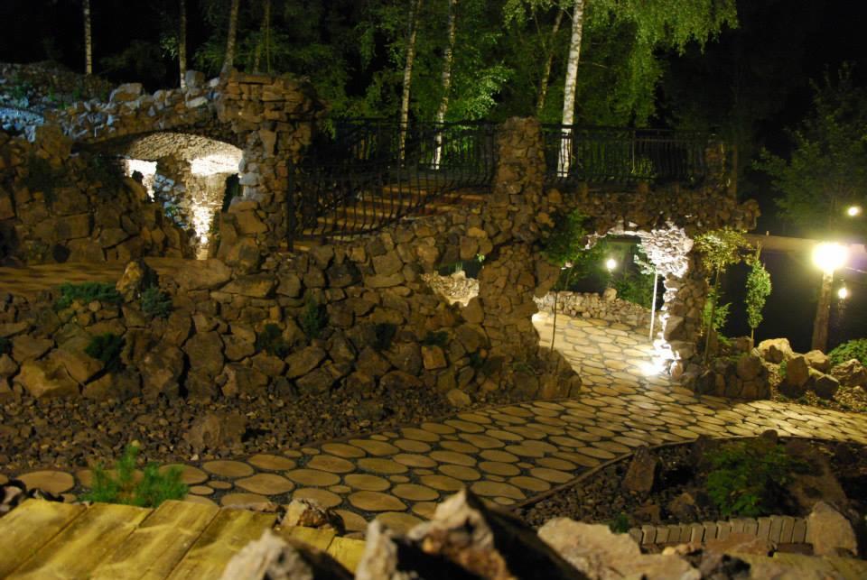 Oświetlony nocą ogród skalny z kamiennym mostkiem, schodami i ścieżką z drewnianych krążków. Dekoracyjne oświetlenie punktowe podkreśla unikalny krajobraz.