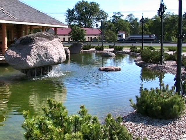 Oczko wodne z dużym głazem pełniącym funkcję fontanny, otoczone roślinnością i oświetlone latarniami, tworzące malowniczy krajobraz.