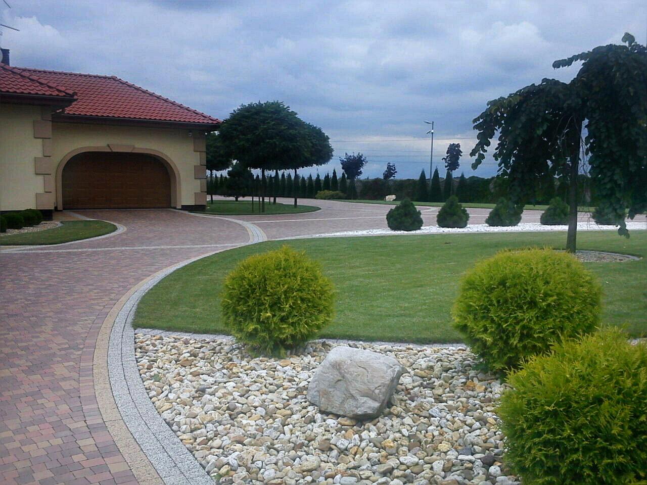 Uporządkowany ogród z kulistymi tujami, kamieniami ozdobnymi i brukowaną ścieżką prowadzącą do domu z garażem. Trawa równo skoszona, drzewa zadbane.