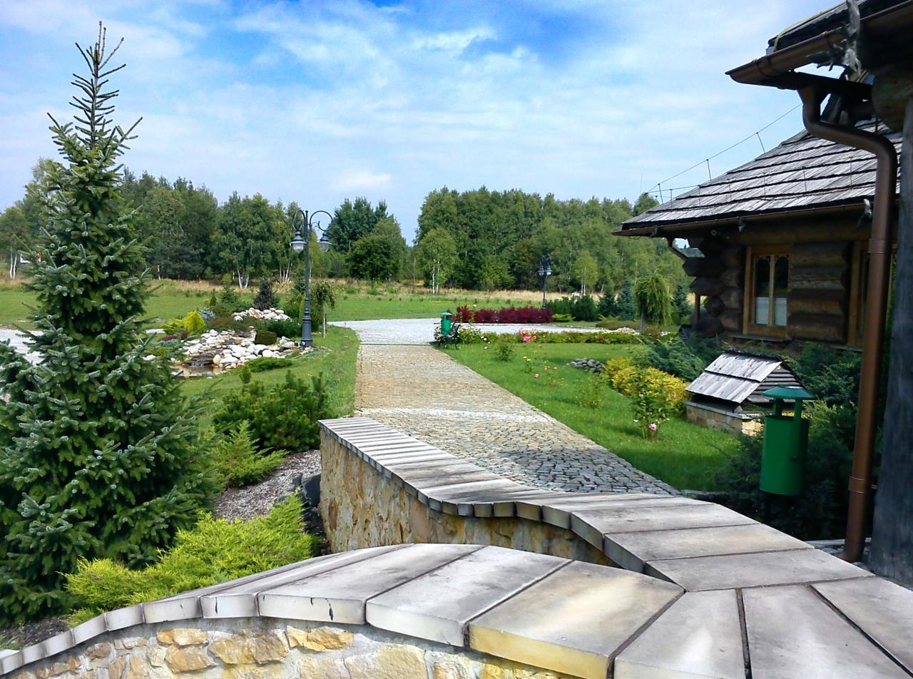 Ogród z kamienną ścieżką, oświetlony lampami, prowadzącą do drewnianego domu. W tle widoczne drzewa i niebo. Elementy małej architektury ogrodowej.