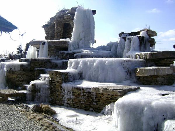 Kamienna kaskada wodna z lodem i śniegiem w słoneczny dzień. Widoczne warstwy kamieni i zamarznięta woda tworzą zimowy krajobraz.