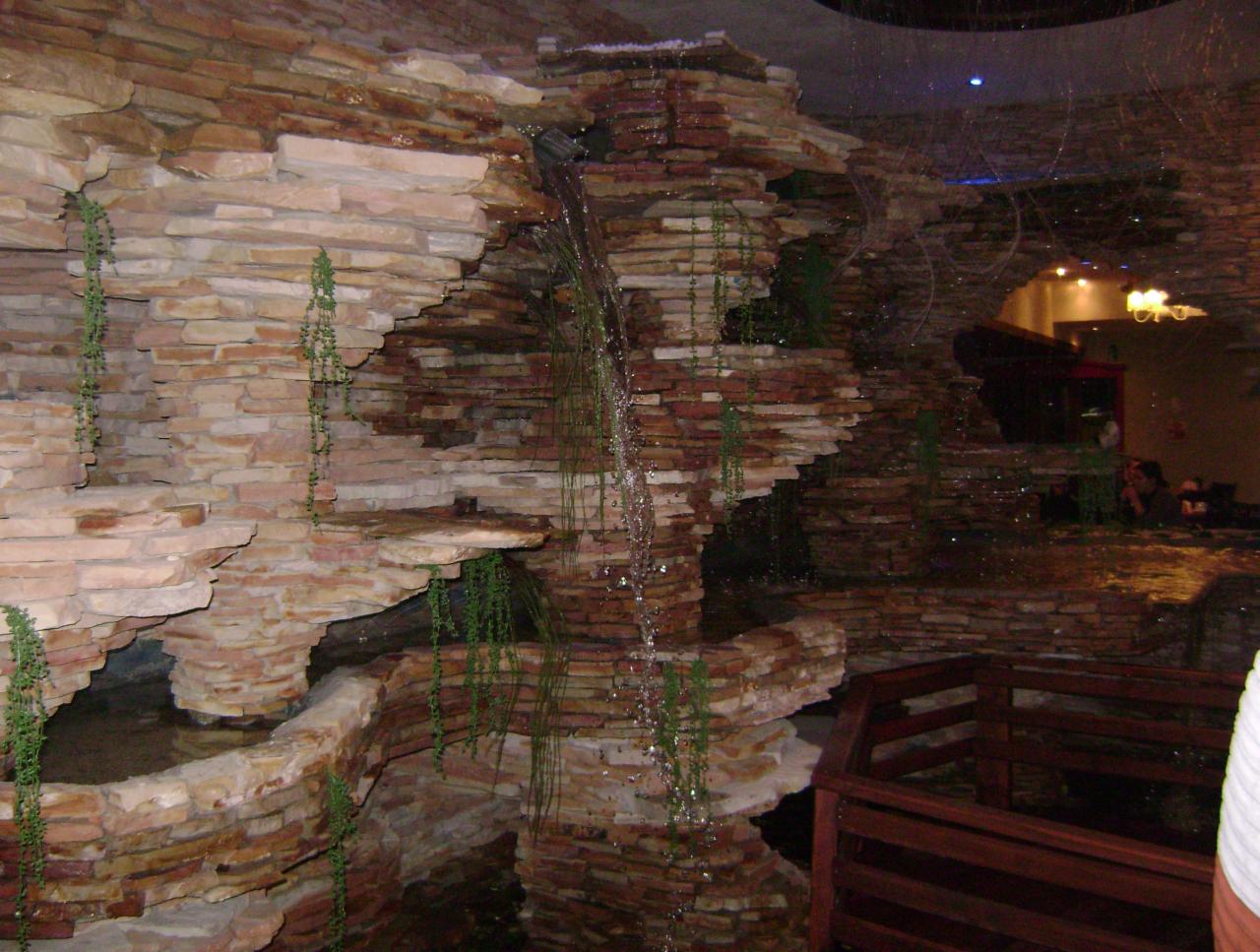 Dekoracyjna ściana z kamienia z kaskadą wodną i zwisającymi roślinami, oświetlona punktowo. Widoczne elementy drewniane i fragment wnętrza z oświetleniem.