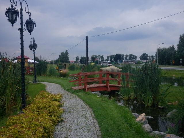 Oświetlony park z kamienną ścieżką, mostkiem i rzeźbą żubra w tle, oświetlony latarniami. Zielona trawa i roślinność. Pochmurne niebo.