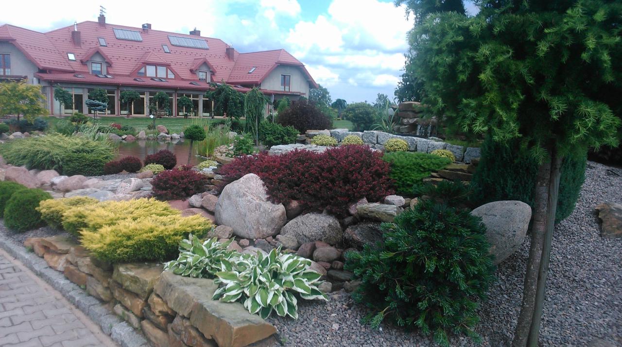 Ogród z oczkiem wodnym i kaskadą, kamiennymi murkami oraz różnorodną roślinnością ozdobną na tle dużego domu z czerwonym dachem i solarnymi panelami.