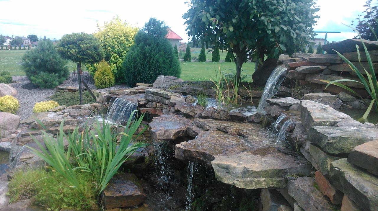 Ogród z kaskadą wodną zbudowaną z kamieni, otoczony zielenią, krzewami i drzewami. Woda spływa po kamieniach tworząc mały wodospad.