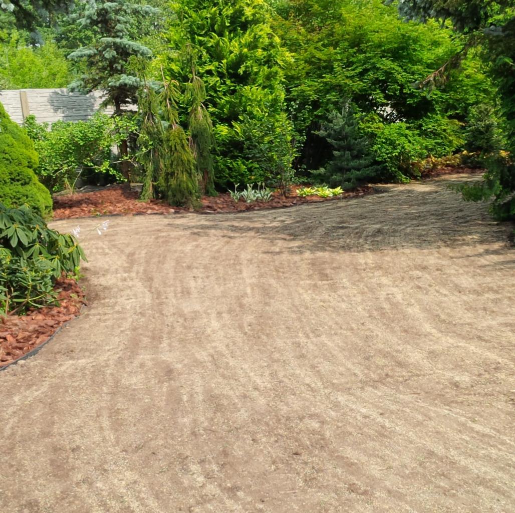 Przygotowany teren pod trawnik z rolki, obrzeża wykończone korą. Widoczne drzewa i krzewy w tle. Grunt wyrównany, gotowy do ułożenia trawy.