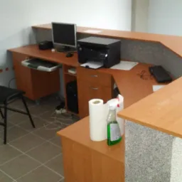 Nowoczesna recepcja z blatem z jasnego drewna i szarym, teksturowanym frontem, wyposażona w komputer, drukarkę i czarne krzesło na tle białej ściany i kafelkowej podłogi. Na blacie widoczny płyn...