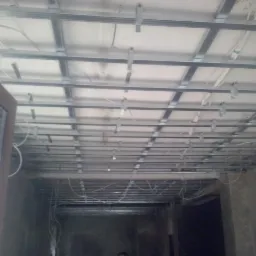 Metalowa konstrukcja sufitu podwieszanego z widocznymi przewodami elektrycznymi, częściowo zamontowana płyta karton-gips, widok od dołu z perspektywy pomieszczenia.