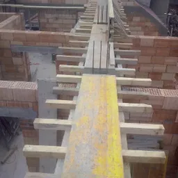 Wznoszenie murów domu z pustaków ceramicznych, widoczne drewniane szalunki i zbrojenie stropu, na drugim planie budynek sąsiadujący z czerwoną dachówką.