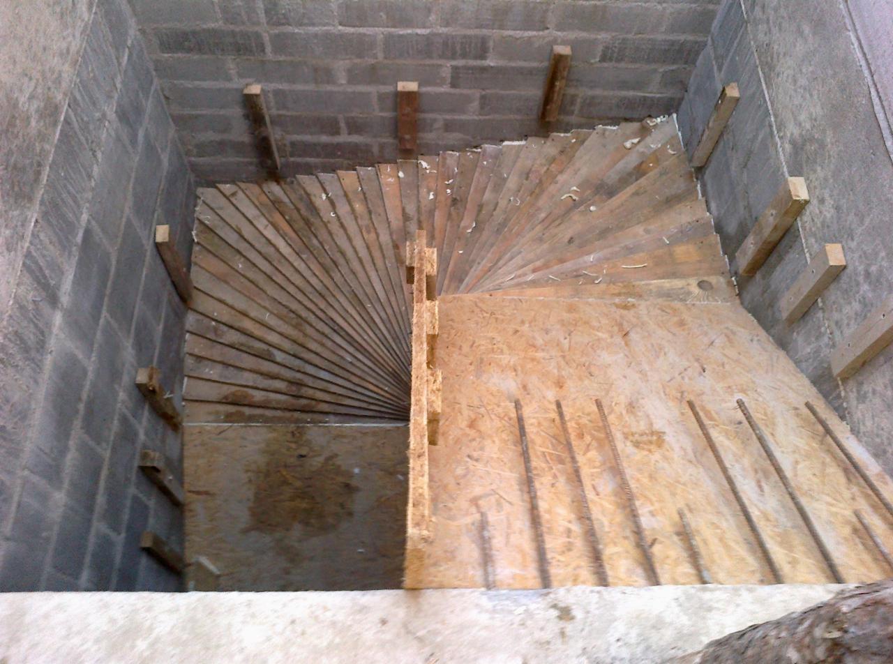 Szare ściany z betonu komórkowego otaczają drewniany szalunek spiralnych schodów z wystającymi prętami zbrojeniowymi, widok z góry.