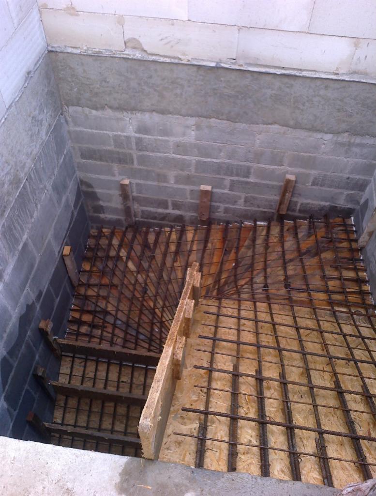Zbrojenie schodów w surowym stanie, widok z góry na konstrukcję z prętów i szalunku z płyty OSB, ściany z szarych bloczków betonowych.