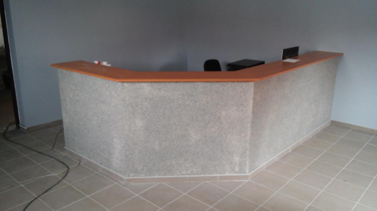 Nowoczesna lada recepcyjna z blatem w kolorze drewna, wykończona szarą, ziarnistą fakturą, w pomieszczeniu w Małomicach z szarymi ścianami i beżową podłogą z płytek. Na blacie widoczny laptop...