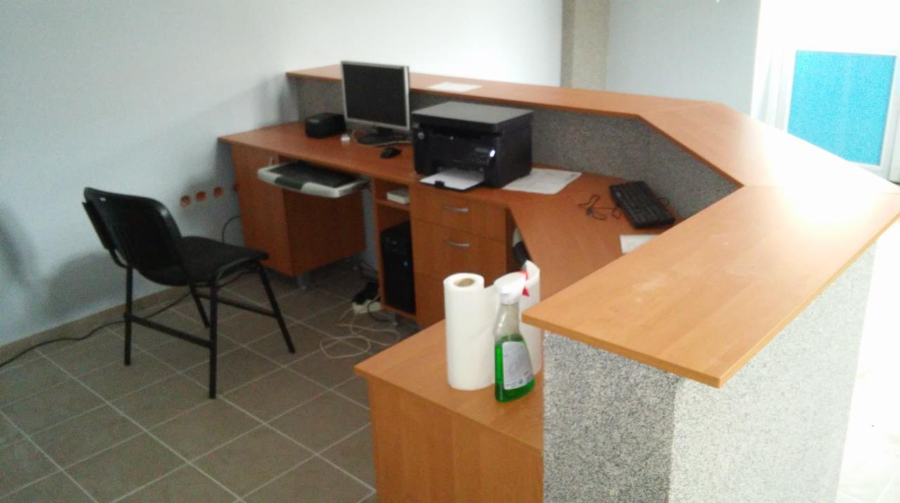 Nowoczesna recepcja z blatem z jasnego drewna i szarym, teksturowanym frontem, wyposażona w komputer, drukarkę i czarne krzesło na tle białej ściany i kafelkowej podłogi. Na blacie widoczny płyn...