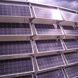 Elewacja budynku pokryta panelami słonecznymi, widoczne mocowania, pochmurne niebo odbija się w panelach.