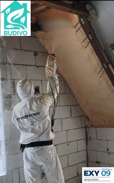 Pracownik w kombinezonie ochronnym aplikuje piankę PUR na poddasze, widoczne krokwie i ocieplona powierzchnia. Na ścianie z bloczków logo firmy i systemu natryskowego.