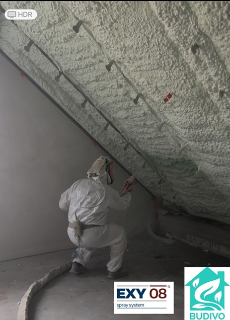 Pracownik w kombinezonie natryskuje pianę PUR na skośny sufit poddasza, widoczne elementy konstrukcyjne. Aplikacja pianki, ocieplenie natryskowe.