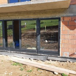 Firma handlowo-usługowa Ewa Korzeniak - Czteroskrzydłowe okno z czarnymi ramami w surowym stanie budynku, widoczne cegły, betonowy strop oraz ziemia przed oknem z fragmentami drewna.