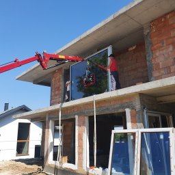 Firma handlowo-usługowa Ewa Korzeniak - Montaż dużej szyby okiennej na budowie za pomocą czerwonego dźwigu przyssawkowego, widoczne dwie osoby w trakcie pracy, w tle surowy stan budynku i błękitne niebo.