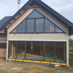 Firma handlowo-usługowa Ewa Korzeniak - Surowy stan budynku z czarnymi oknami aluminiowymi, widoczne cegły i beton, żółte elementy wykończeniowe przy gruncie, maszt telekomunikacyjny w tle.