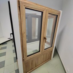 Firma handlowo-usługowa Ewa Korzeniak - Dwu-skrzydłowe okno z drewnopodobną ramą, widoczne z boku, z uchwytem w kolorze mosiądzu, eksponowane w jasnym pomieszczeniu na tle innych okien.