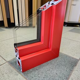 Firma handlowo-usługowa Ewa Korzeniak - Przekrój poprzeczny czerwonej ramy okiennej z trzema szybami, ukazujący wewnętrzną konstrukcję profilu, na tle podłogi z płytek i fragmentu ekspozycji drzwi.