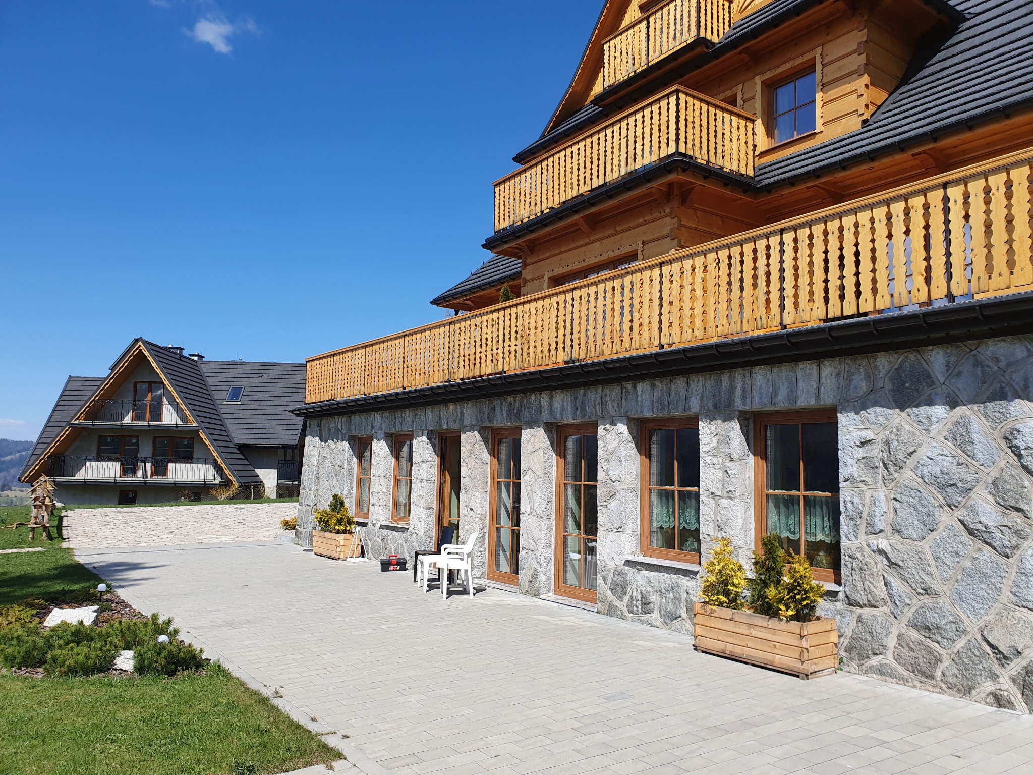 Drewniane okna w domu w stylu góralskim, widoczne z zewnątrz, z kamienną elewacją i drewnianymi ramami, z dekoracyjnymi firankami w oknach, na tle błękitnego nieba i drugiego budynku.