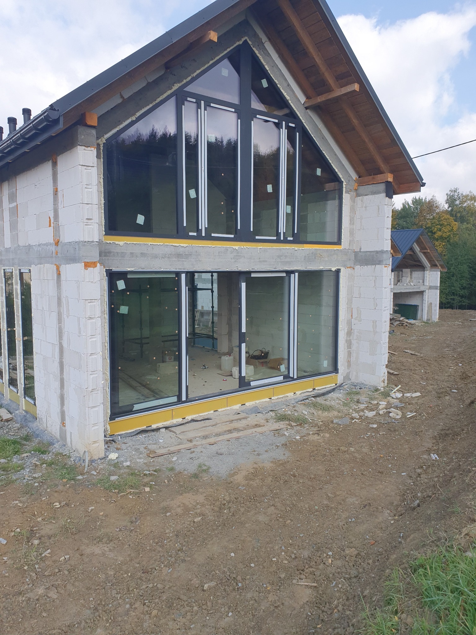 Surowy stan domu z bloczków z zamontowanymi dużymi oknami w czarnych ramach, widoczne zielone naklejki na szybach i żółta izolacja pod oknami, drugi budynek w tle.