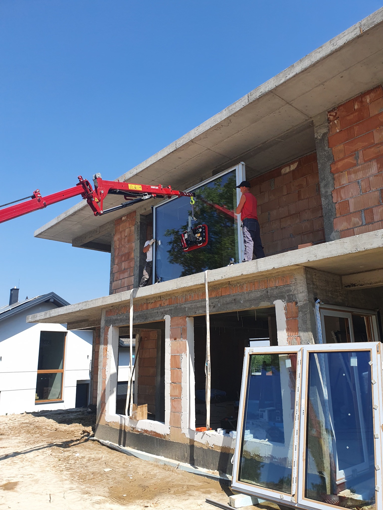 Montaż dużej szyby okiennej na budowie za pomocą czerwonego dźwigu przyssawkowego, widoczne dwie osoby w trakcie pracy, w tle surowy stan budynku i błękitne niebo.