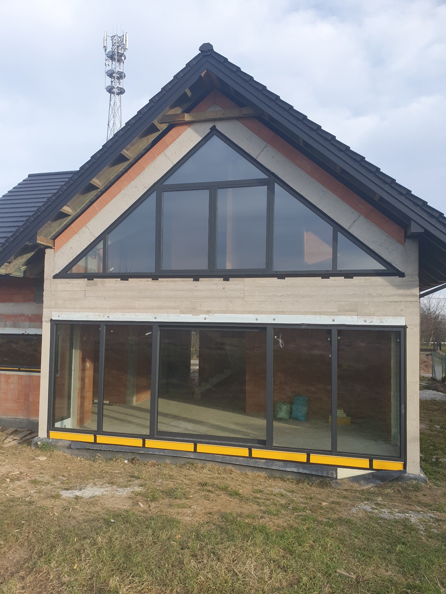Surowy stan budynku z czarnymi oknami aluminiowymi, widoczne cegły i beton, żółte elementy wykończeniowe przy gruncie, maszt telekomunikacyjny w tle.
