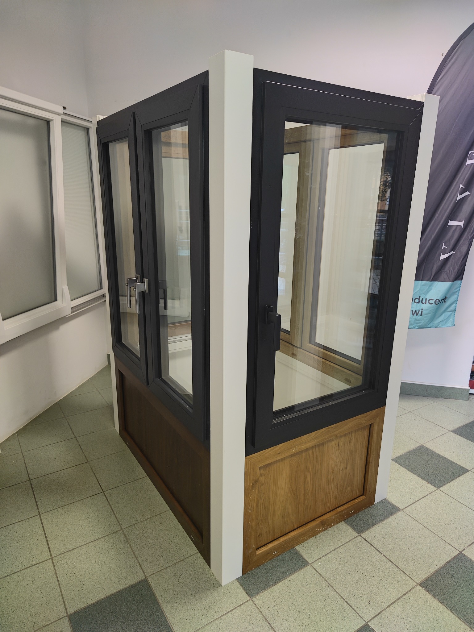 Ekspozycja narożna z czarnymi ramami okiennymi i drewnianą podstawą, prezentowana w salonie z otwartym oknem w tle.