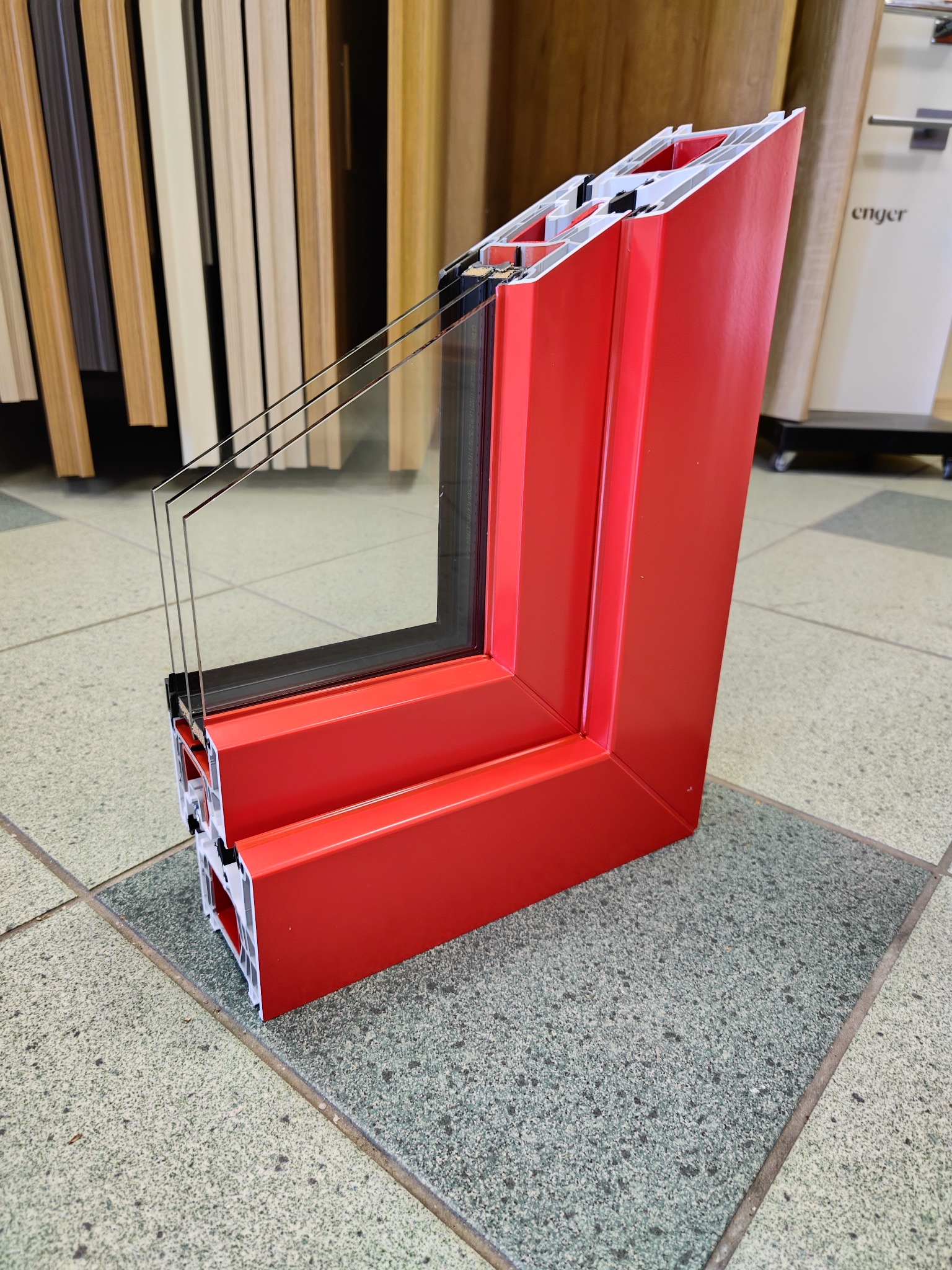 Przekrój poprzeczny czerwonej ramy okiennej z trzema szybami, ukazujący wewnętrzną konstrukcję profilu, na tle podłogi z płytek i fragmentu ekspozycji drzwi.