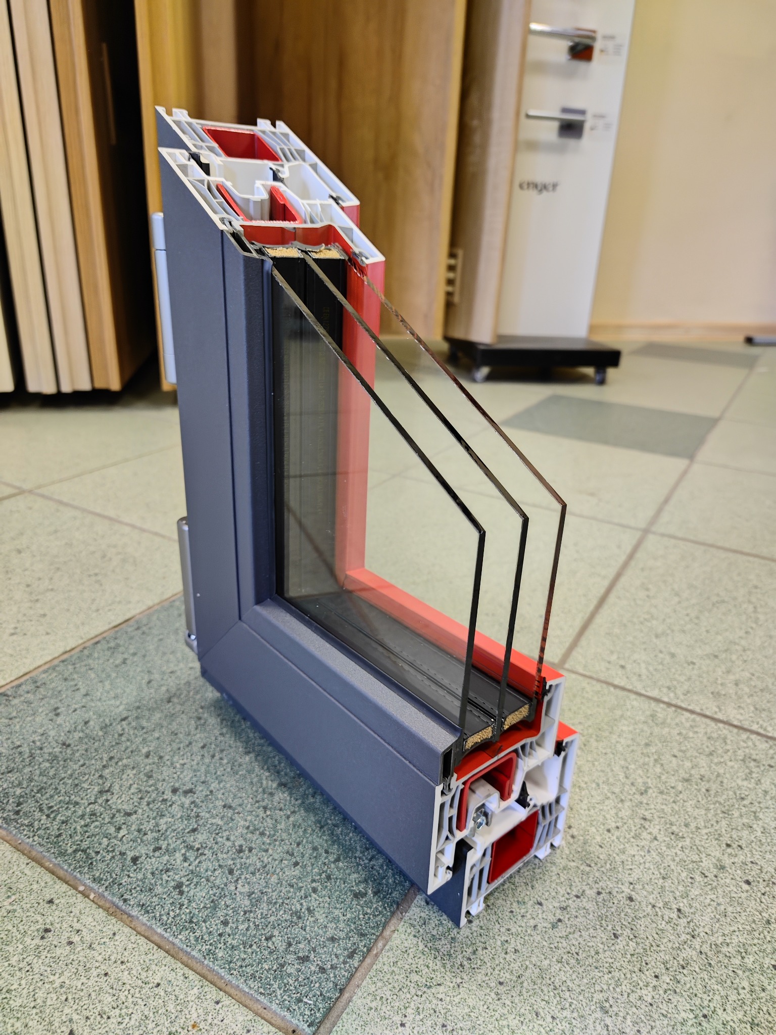 Przekrój okna z trzema szybami w szarej ramie, prezentujący budowę i izolację, na tle płytek podłogowych w Krakowie.