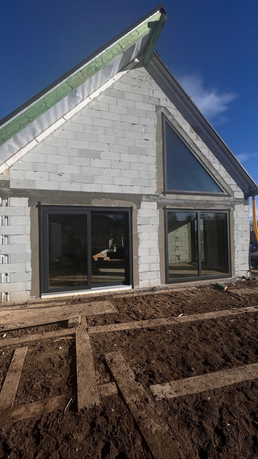 Surowy stan budynku z szarego bloczka z zamontowanymi czarnymi oknami przesuwnymi i trójkątnym oknem na szczycie, widoczne deski na ziemi.