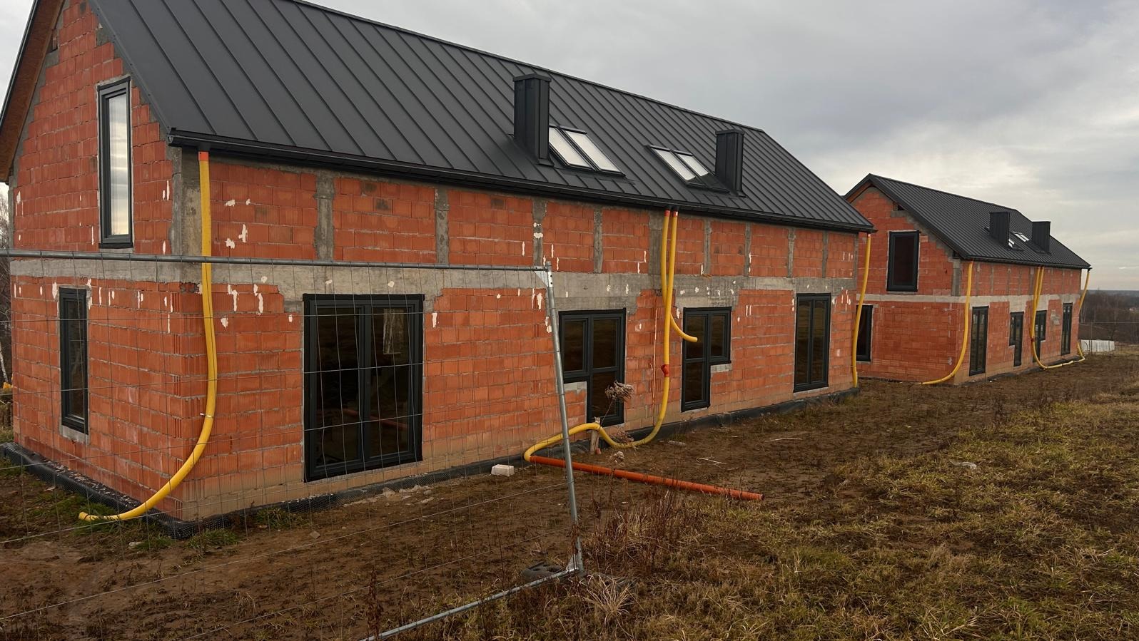 Surowy stan dwóch budynków z czerwonej cegły z czarnymi ramami okiennymi i czarnym dachem, żółte rury wzdłuż ścian, widok z zewnątrz przez ogrodzenie.