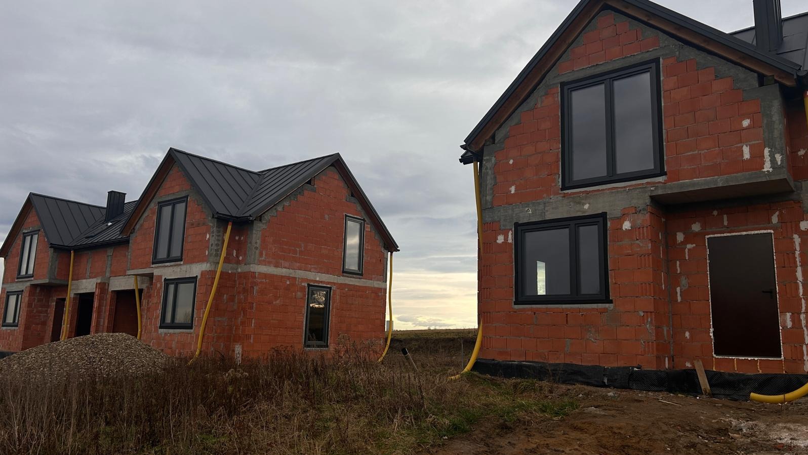 Surowe, ceglane domy w trakcie budowy z zamontowanymi czarnymi oknami i rynnami, widoczne na tle pochmurnego nieba i pola.