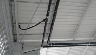 Czarne rury instalacyjne zamocowane do białego sufitu hali, widok z dołu, z fragmentem konstrukcji ściany po lewej.