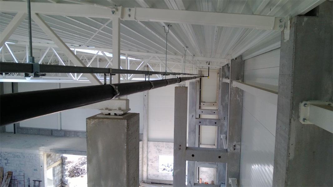 Wnętrze hali produkcyjnej w trakcie budowy, widoczne betonowe słupy konstrukcyjne, stalowa konstrukcja dachu pomalowana na biało oraz rurociąg podwieszony pod sufitem na metalowych wspornikach.