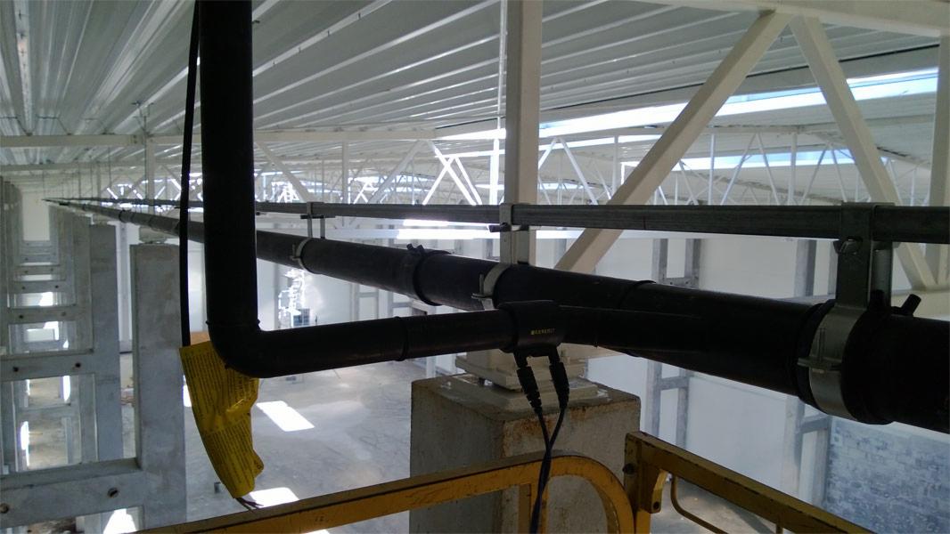 Czarne rury wentylacyjne zamontowane pod sufitem hali przemysłowej, widoczne elementy mocujące i podwieszana konstrukcja sufitu.