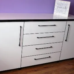 Biała komoda z czterema szufladami i szafkami, czarne uchwyty, czarny blat, oświetlona dekoracja z napisem 'KEEP LIFE SIMPLE & SMILE' na fioletowym tle ściany.