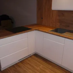 Kąt ujęcia na fragment białej, nowoczesnej kuchni z drewnianym blatem, w tle karton z logo Samsung, widoczna płyta indukcyjna i zlew.