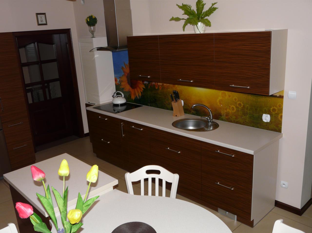 Nowoczesna kuchnia z brązowymi szafkami z poziomymi paskami, blatem w jasnym odcieniu i panelem szklanym z motywem słoneczników, uzupełniona okrągłym stołem z bukietem tulipanów.