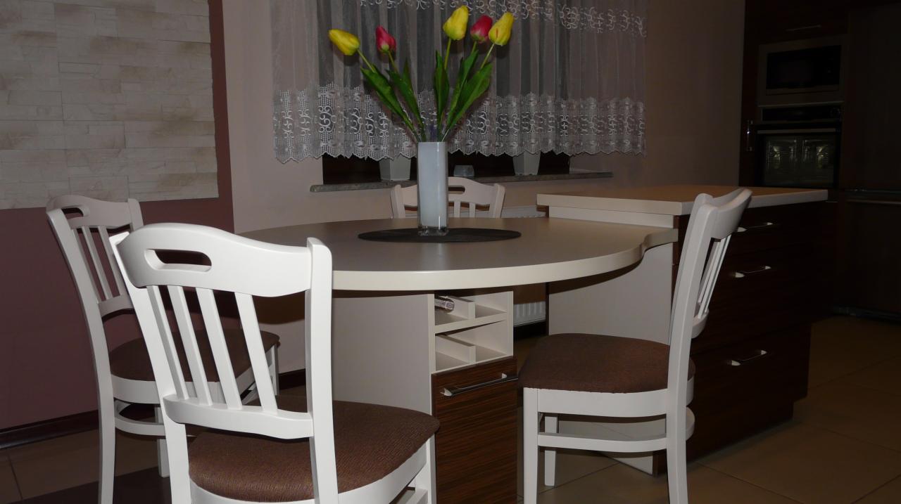 Okrągły stół z białym blatem i wbudowanym barkiem, otoczony białymi krzesłami z brązowymi siedziskami, w tle zabudowa kuchenna z piekarnikiem i mikrofalówką.