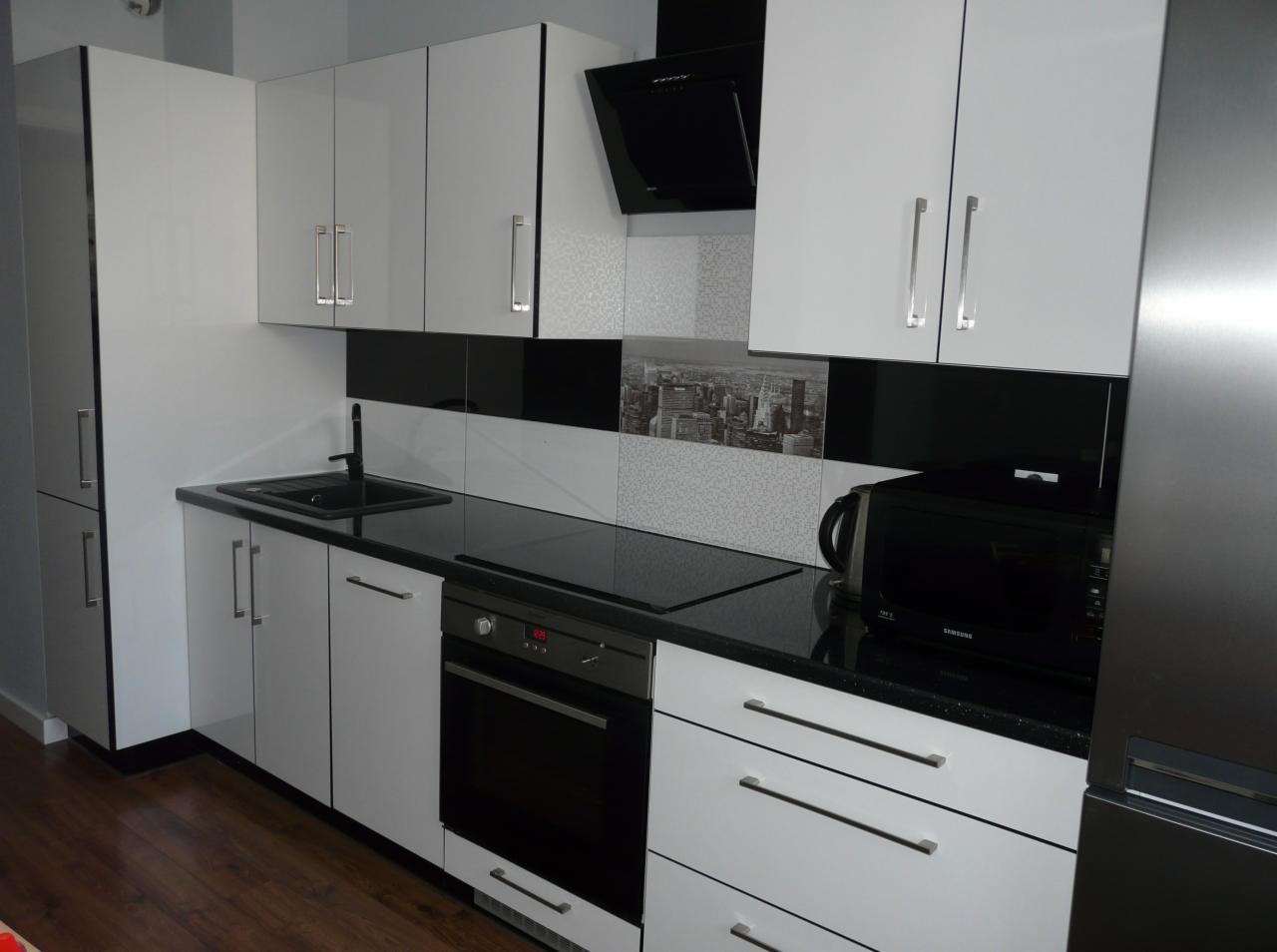 Nowoczesna kuchnia z białymi szafkami, czarnym blatem i płytkami z motywem miejskim, czarny zlewozmywak i piekarnik w zabudowie.