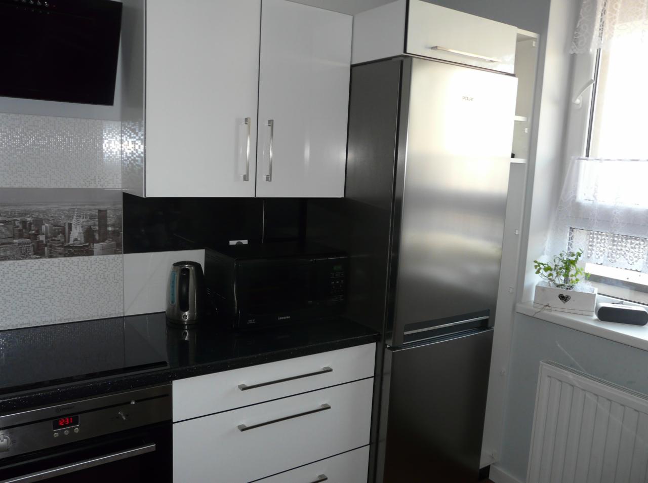 Nowoczesna kuchnia z białymi szafkami, czarnym blatem, srebrną lodówką i czajnikiem elektrycznym na tle ściany z motywem miasta.