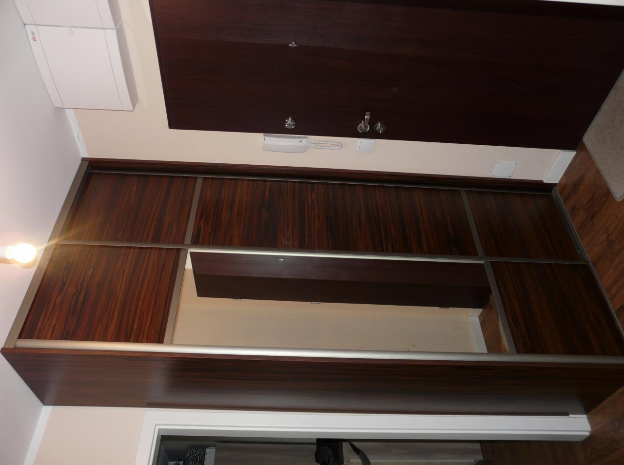 Szafa wnękowa z przesuwnymi drzwiami w kolorze ciemnego drewna z widocznymi metalowymi prowadnicami, częściowo otwarta.