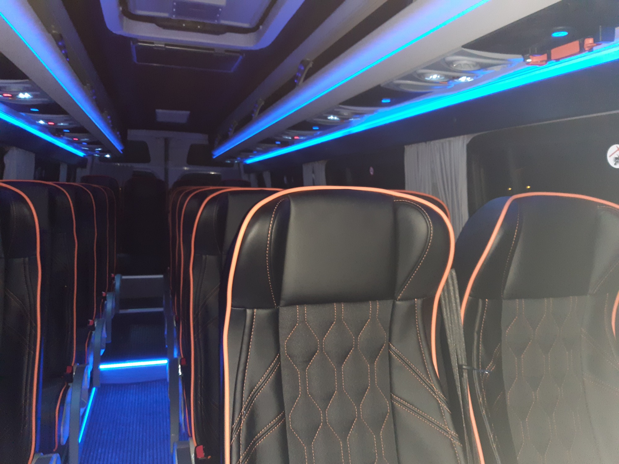 Wnętrze autobusu z czarnymi skórzanymi fotelami obszytymi pomarańczową nicią, oświetlenie sufitowe LED w kolorze niebieskim.