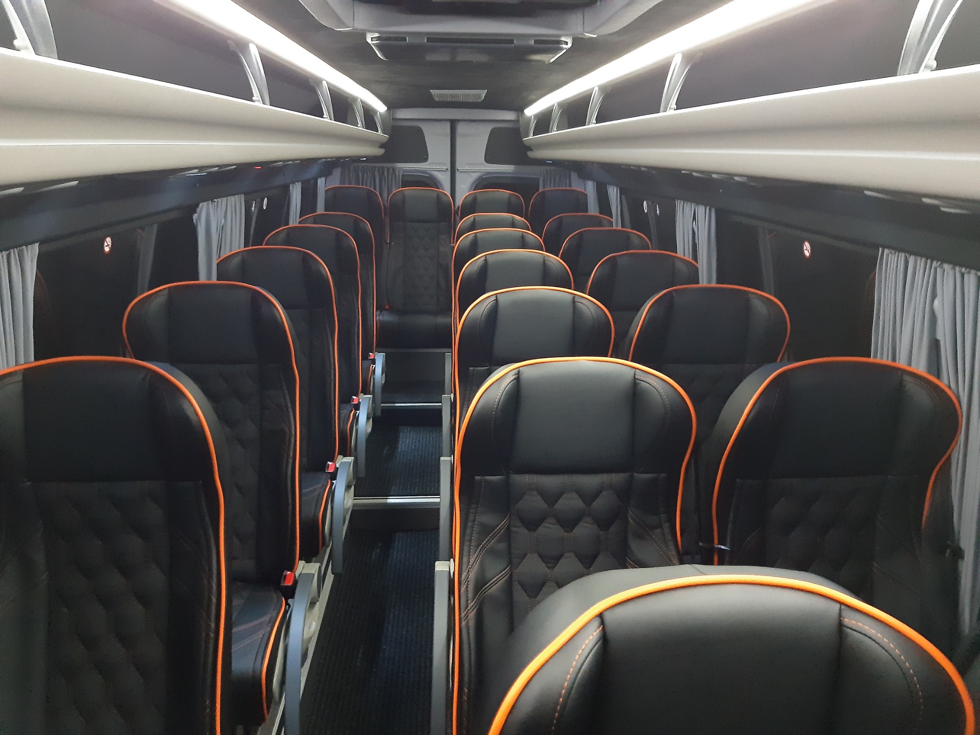 Wnętrze luksusowego busa z czarnymi skórzanymi fotelami z pomarańczowymi przeszyciami, zasłonięte okna, oświetlenie sufitowe.