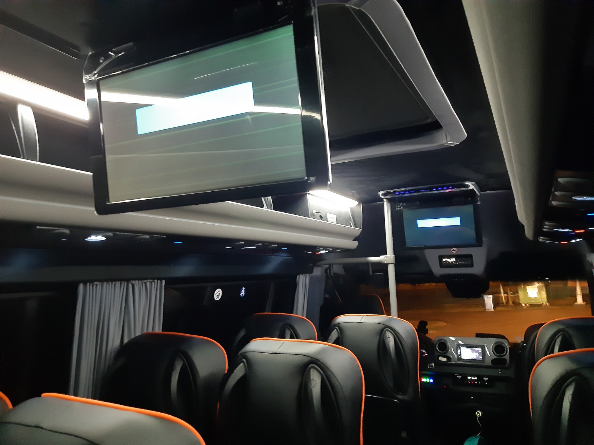 Wnętrze autobusu turystycznego z czarnymi skórzanymi fotelami z pomarańczowym obszyciem i ekranami LCD zamontowanymi pod sufitem.