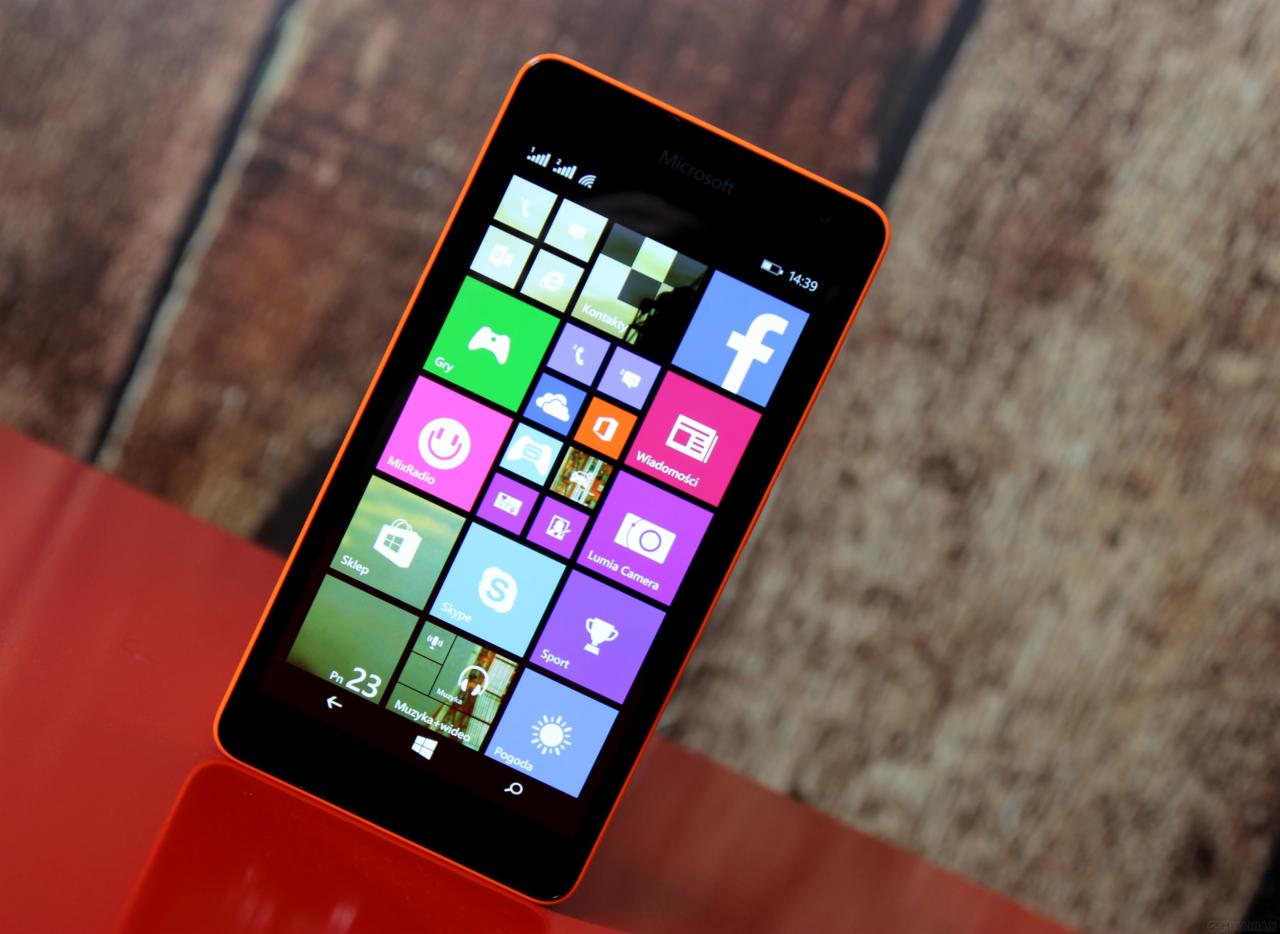 Telefon Microsoft Lumia z pomarańczową obudową, wyświetlający ekran główny z kolorowymi kafelkami aplikacji na tle drewnianego stołu.